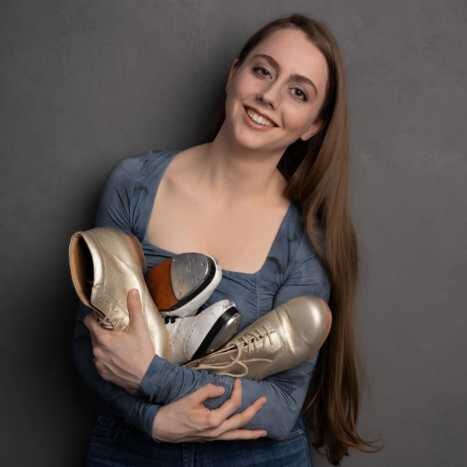Woman wearing blue/gray blouse holding two pairs of tap shoes in gold and white standing in front of a gray wall