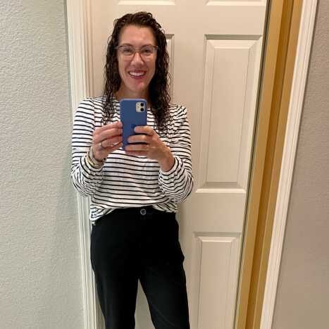 woman in black and white striped long sleeve shirt tucked into black pants. Standing in front of a white door/light gray wall. Holding a mobile phone.